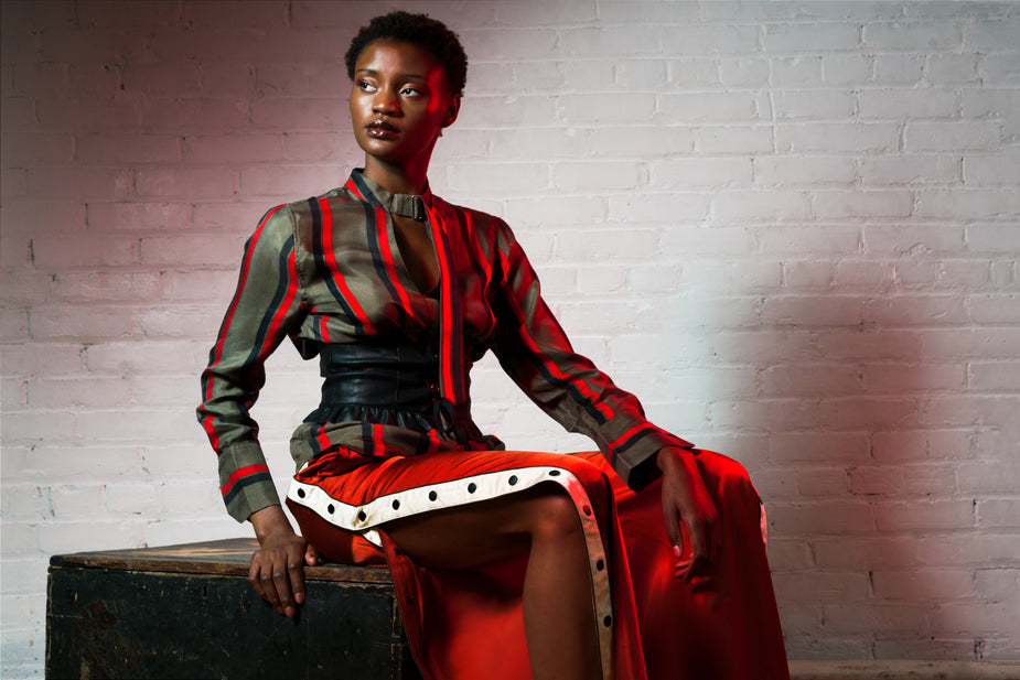 Striped silk blouse in red and olive green with black waist belt, worn by model seated against brick wall.