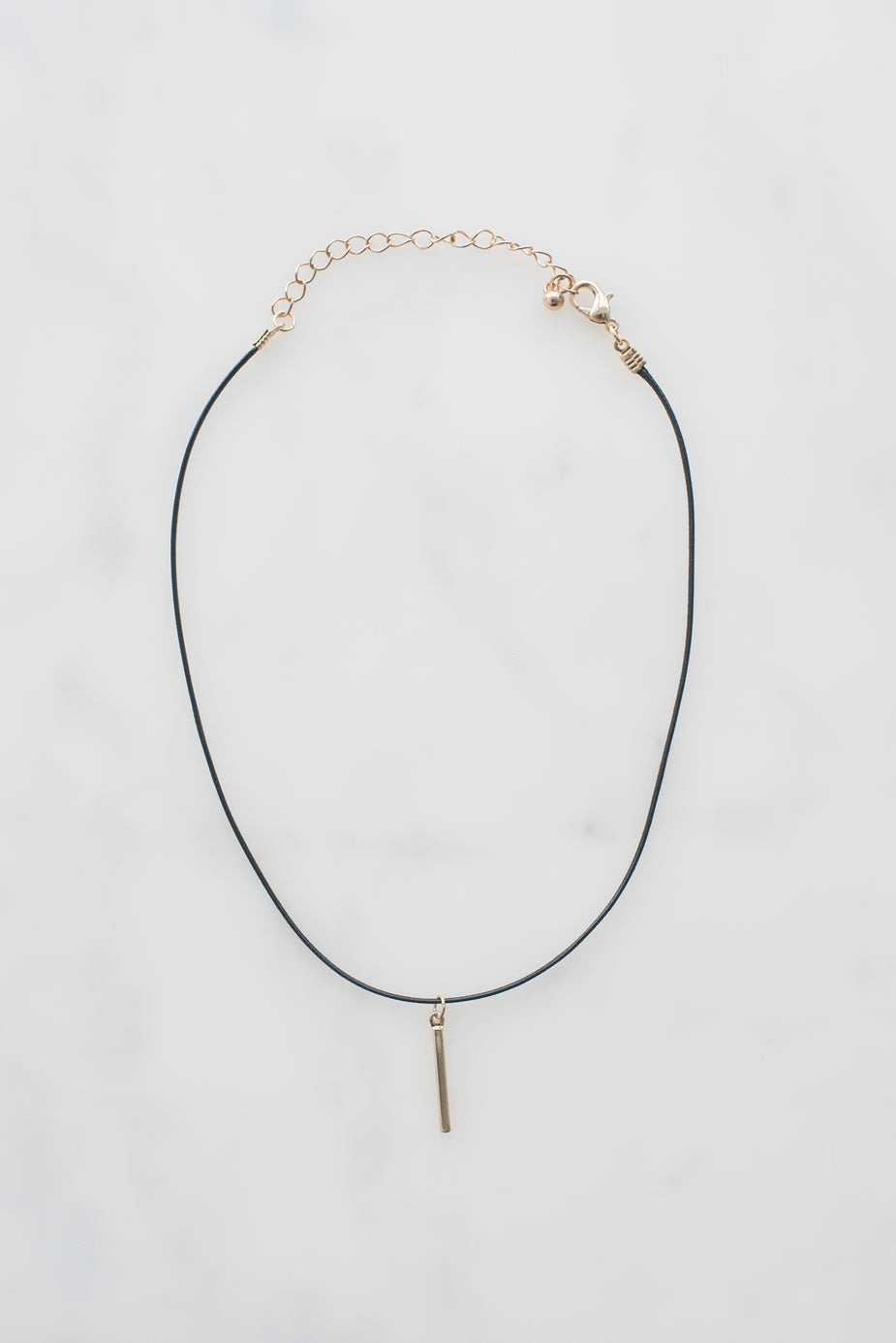 Black leather choker necklace with gold chain extender, lobster clasp, and small gold bar pendant on white background.