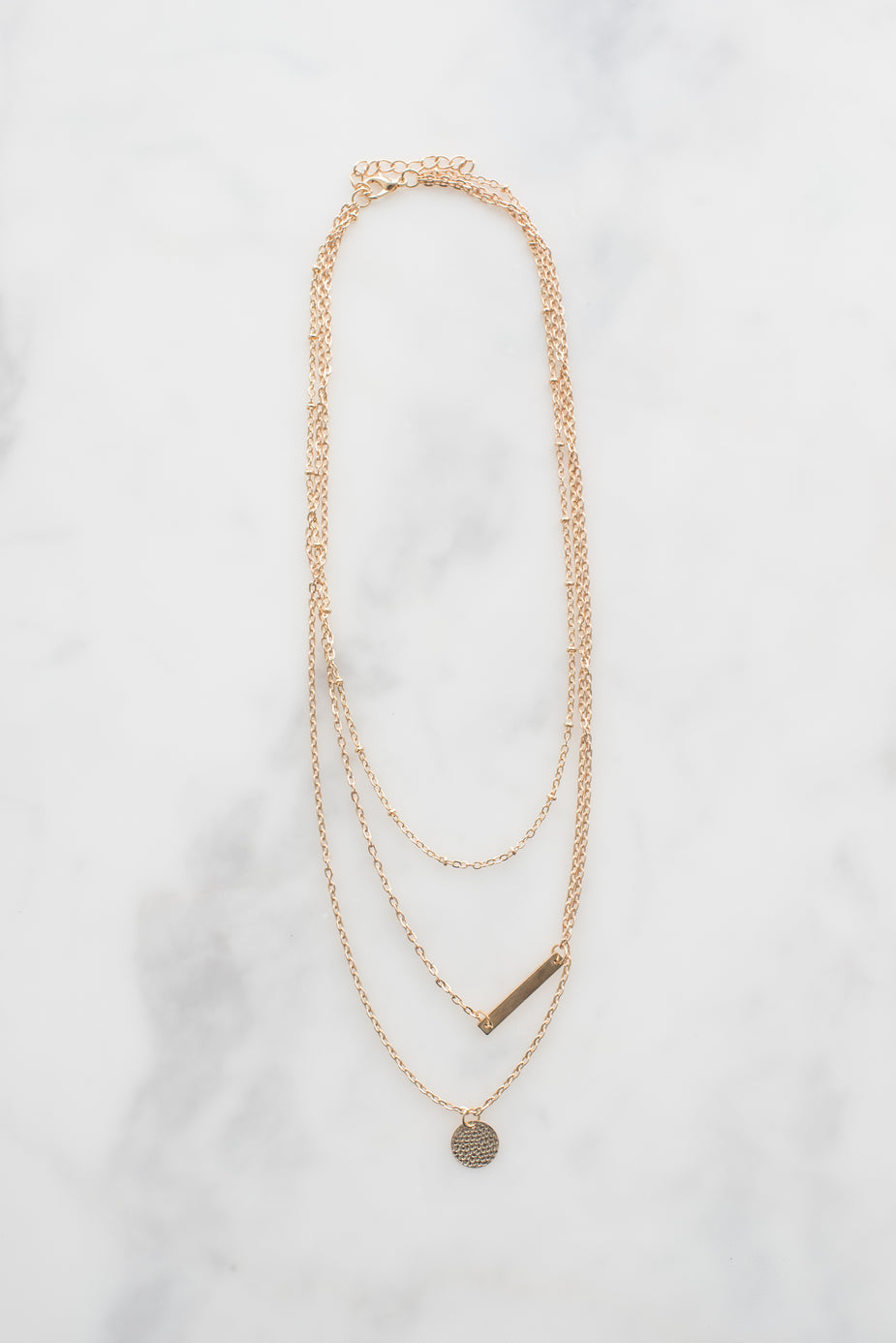 Dainty gold necklace with delicate chain layers, featuring a small disc pendant and bar charm on marble background.