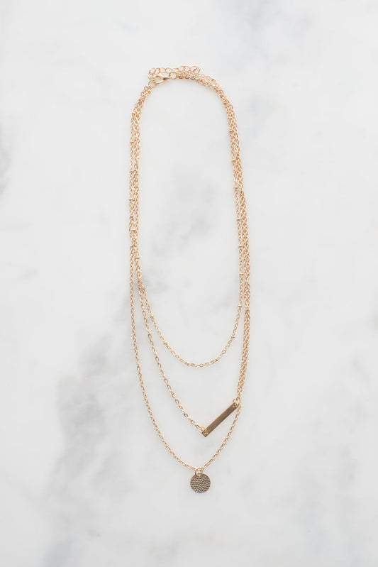 Dainty gold necklace with delicate chain layers, featuring a small disc pendant and bar charm on marble background.