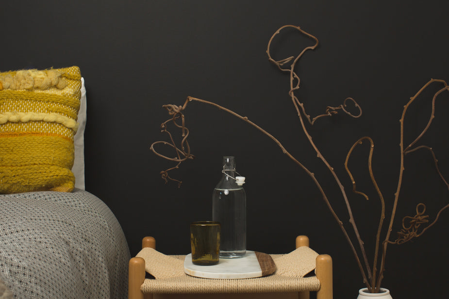 Wooden bedside table with woven seat displaying perfume bottles and styled with dried branches in modern bedroom decor.