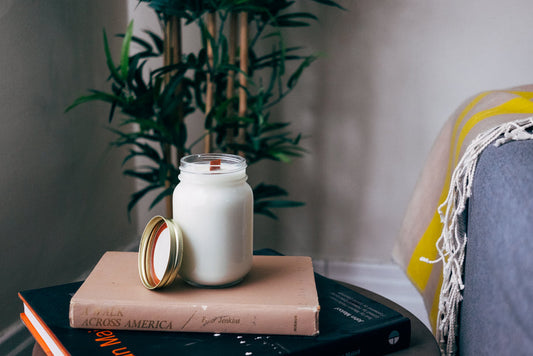Vanilla candle in white glass jar with lid displayed on stacked books, decorated with gold ring, beside plant and yellow pillow