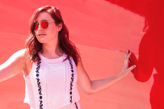 Woman wearing white floral embroidered top with round sunglasses against coral red background