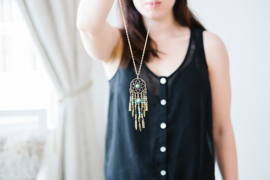 Woman wearing dreamcatcher pendant necklace with turquoise beads and gold chain on black button-up vest.