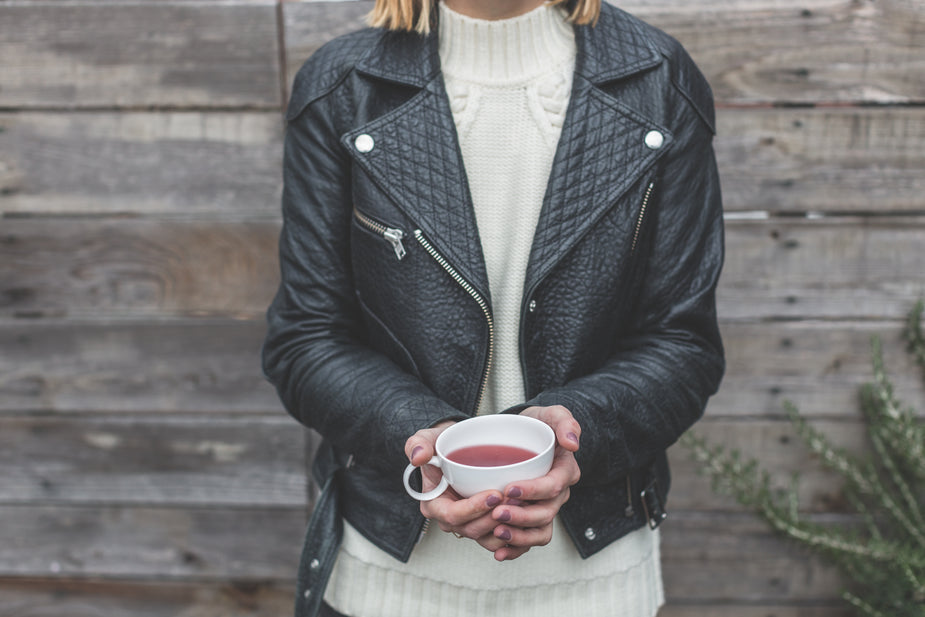 Woman wearing black quilted leather jacket with white sweater, holding warm beverage cup outdoors.
