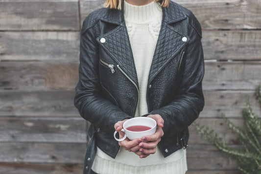 Woman wearing black quilted leather jacket with white sweater, holding warm beverage cup outdoors.