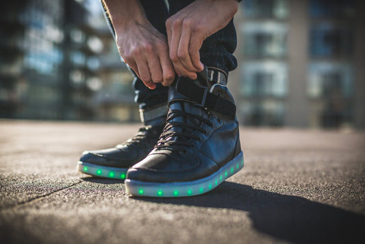 Black LED high tops with glowing green and blue lights on sole, person tying laces outdoors