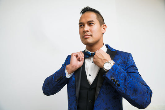Man wearing elegant blue silk tuxedo with black vest, white dress shirt, and blue bow tie against white background.