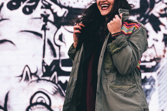 Woman wearing olive green jacket with rainbow stripe trim smiling in front of urban graffiti wall art