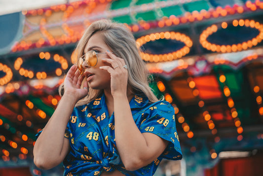 Woman wearing blue silk summer top with number pattern, adjusting yellow sunglasses near illuminated carnival lights