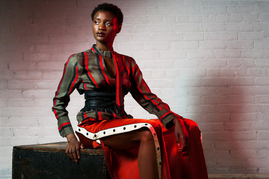 Striped silk blouse in red and olive green with black waist belt, worn by model seated against brick wall.
