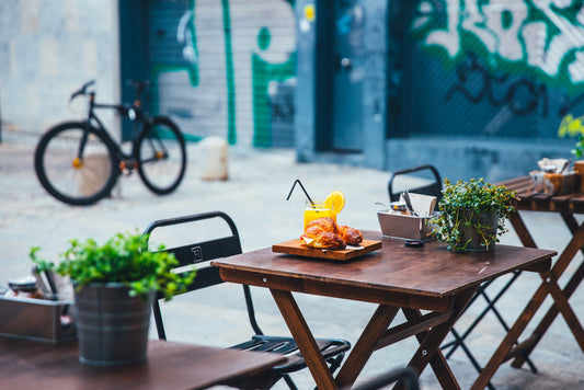 Wooden outdoor table with food and drinks in urban patio setting, potted plants, and bicycle in background