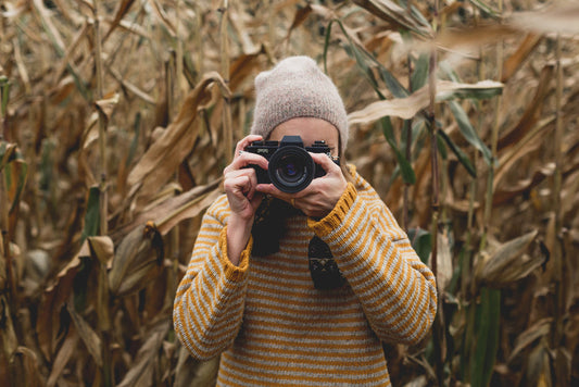 Person wearing yellow wool jumper and beanie holding camera in autumn cornfield